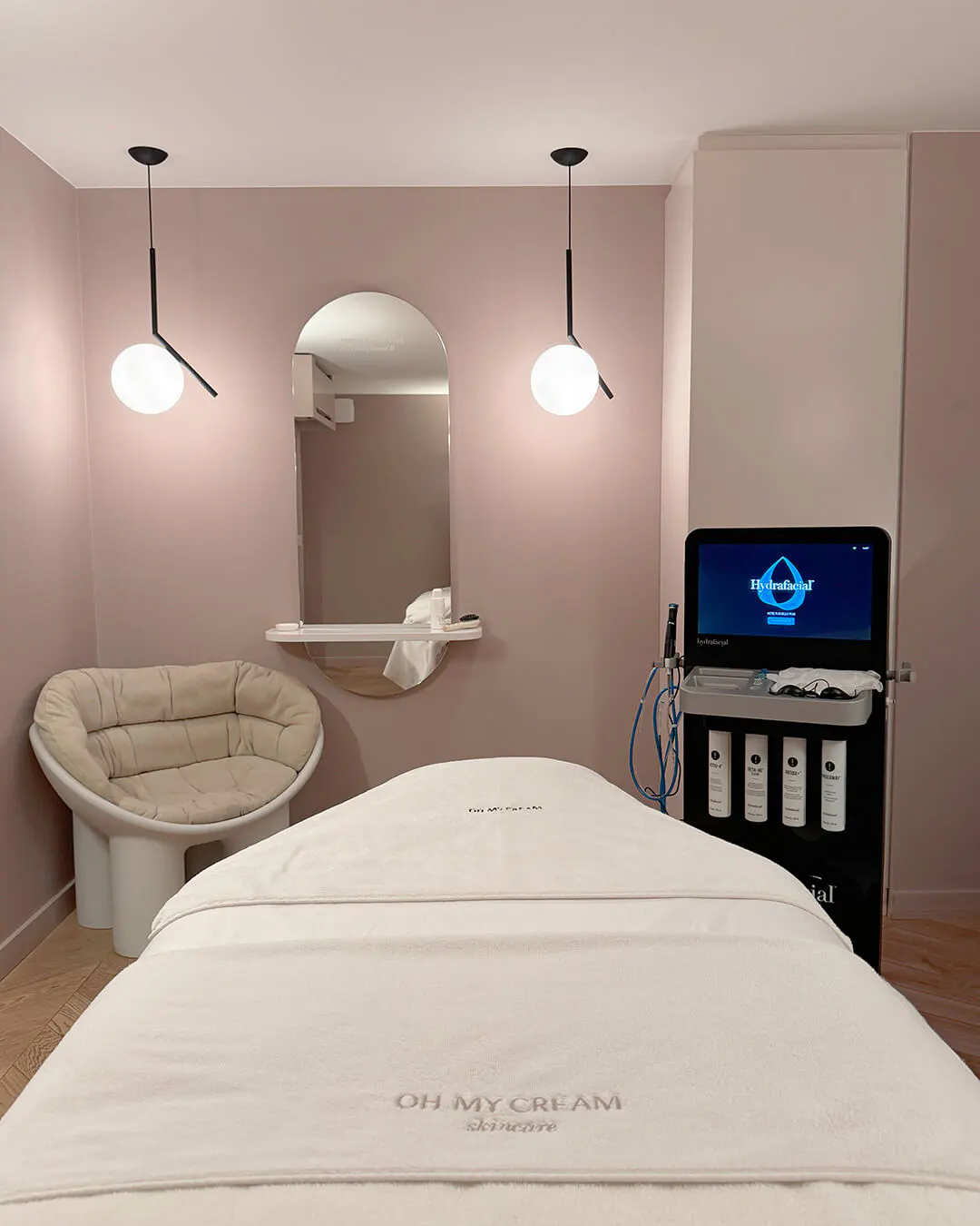 An Oh My Cream treatment cabin: massage table, arched mirror and a facial care device, in a powder-pink room